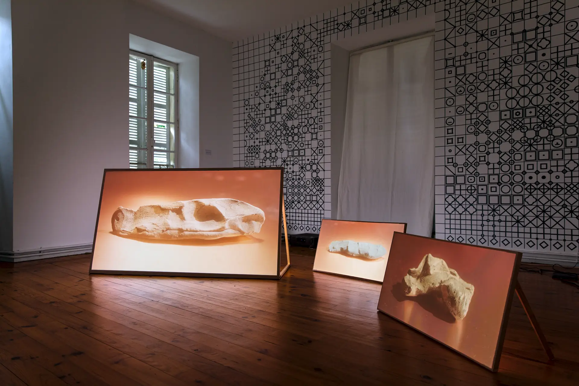 Trois écrans de différentes tailles, posés au sol, diffusent des images d'artefacts en terre sous une lumière chaude, vue d'exposition de l'installation vidéo Manipulations (2025).