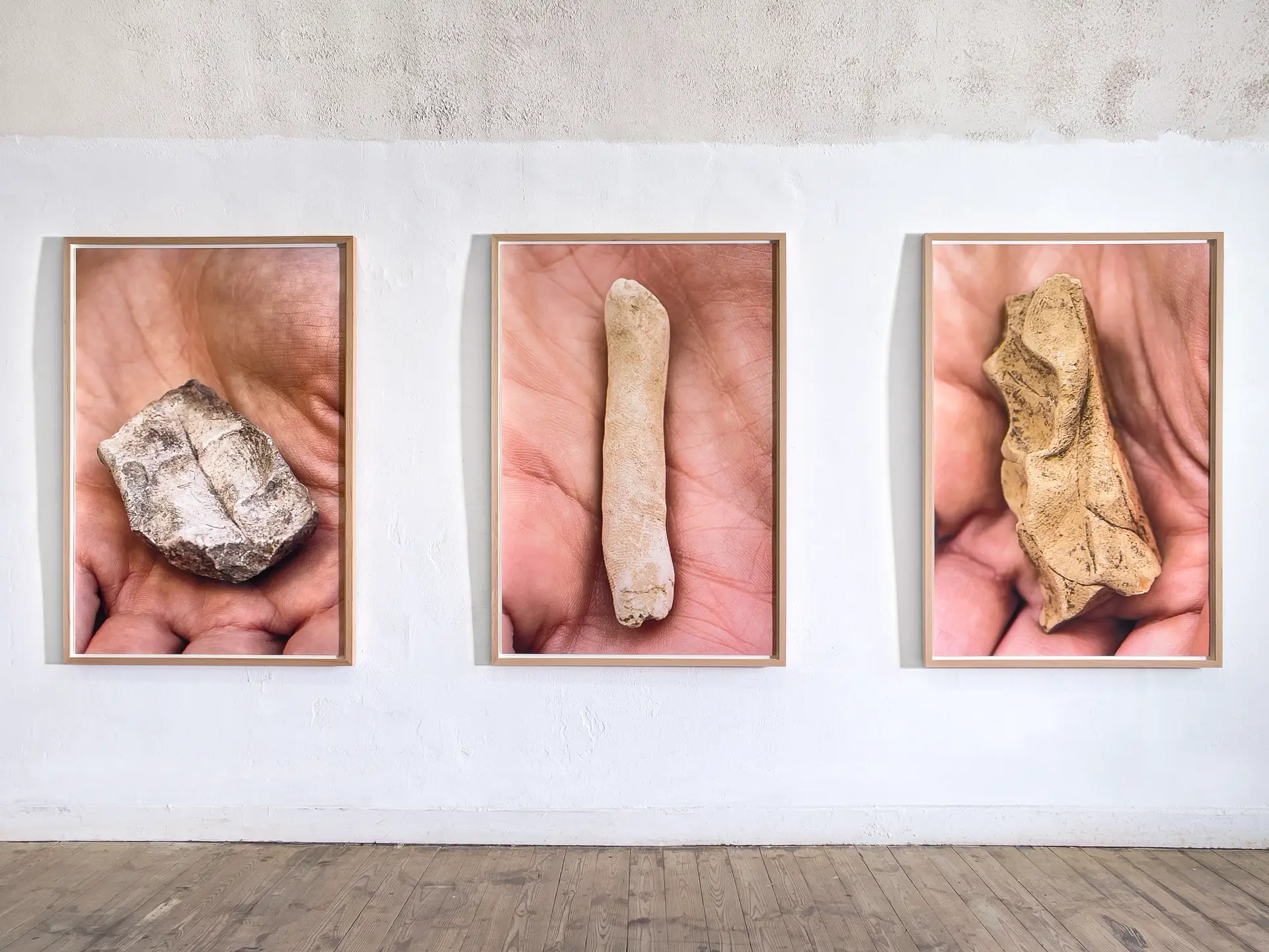 Des artefacts en argile reposent aux creux de mains fortement agrandies. Trois photographies encadrées de bois naturel, vue d'accrochage de la série Muddy Doodle (2024).