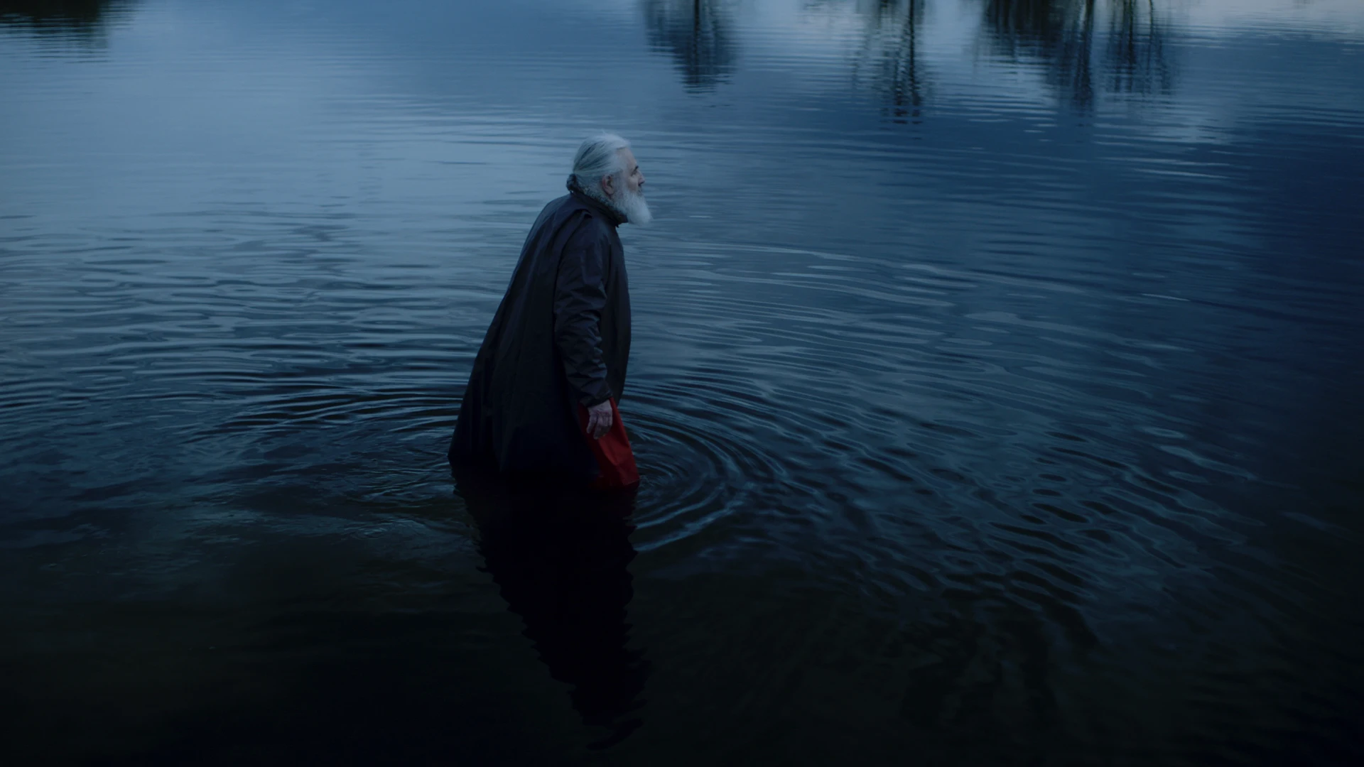Un homme à barbe blanche portant une cape traverse une étendue d'eau sombre, photogramme du triptyque vidéo Translation (2020).